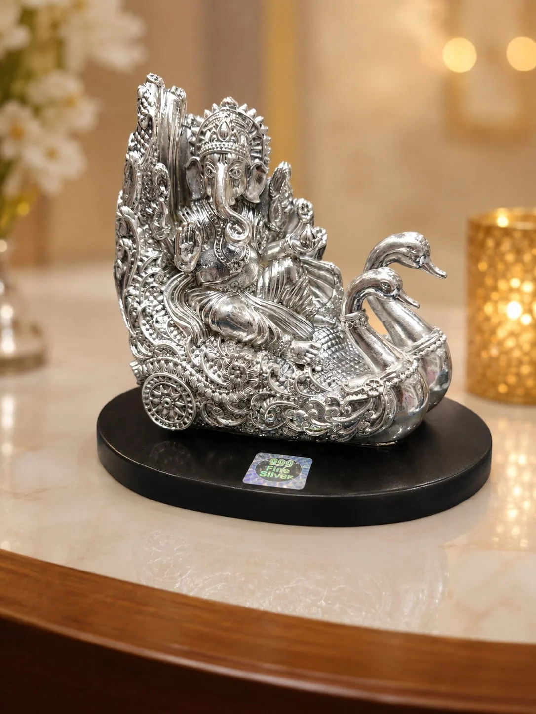 Glossy silver idol of Lord Ganesha sitting on an ornate swan chariot with intricate floral patterns and wheels.