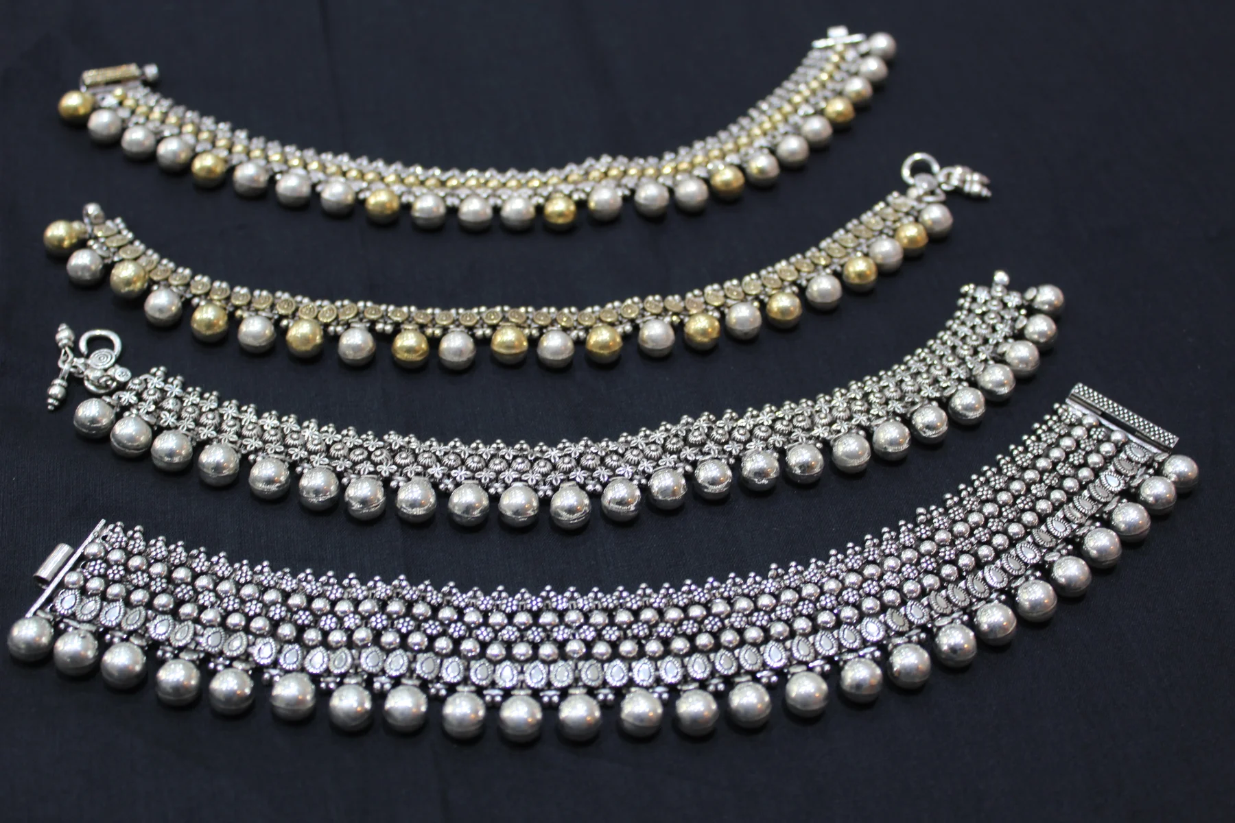 Four silver payal designs featuring intricate patterns and hanging metal beads along the lower edges.