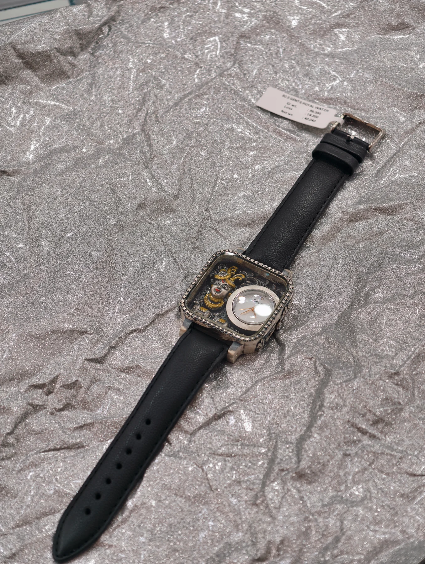 Silver luxury watch with black leather strap, square stone studded dial, and miniature deity face artwork.