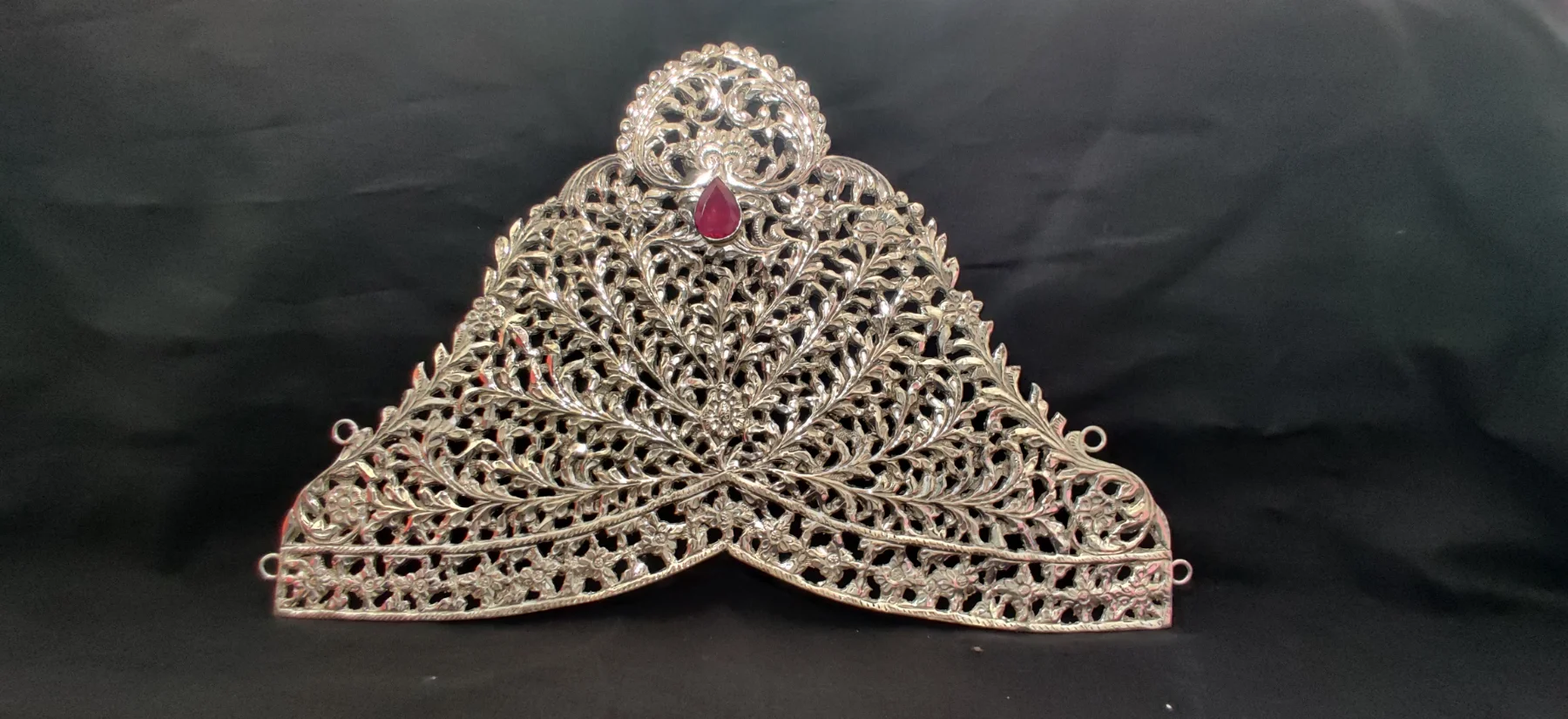 A silver crown features intricate floral filigree work and a central red stone.
