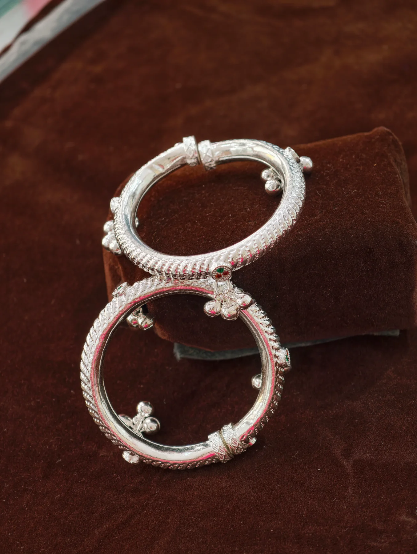 Silver hollow kids kada with textured patterns, small dangling ghungroos, and colourful stones.