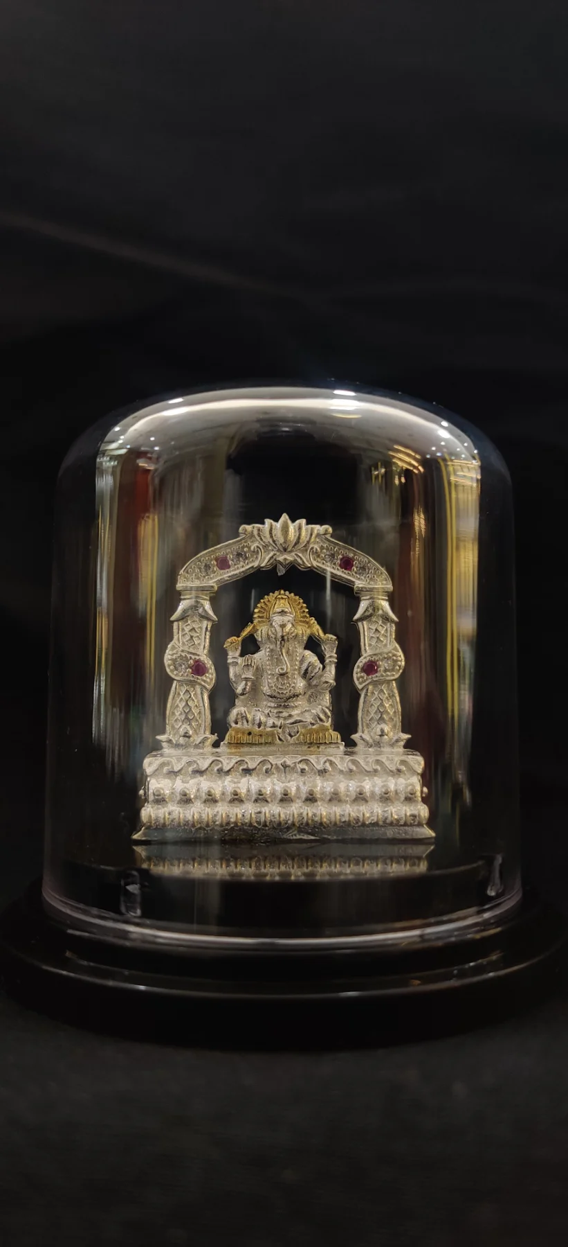 Silver Ganesha idol under a glass dome with ornate arch and red stones.