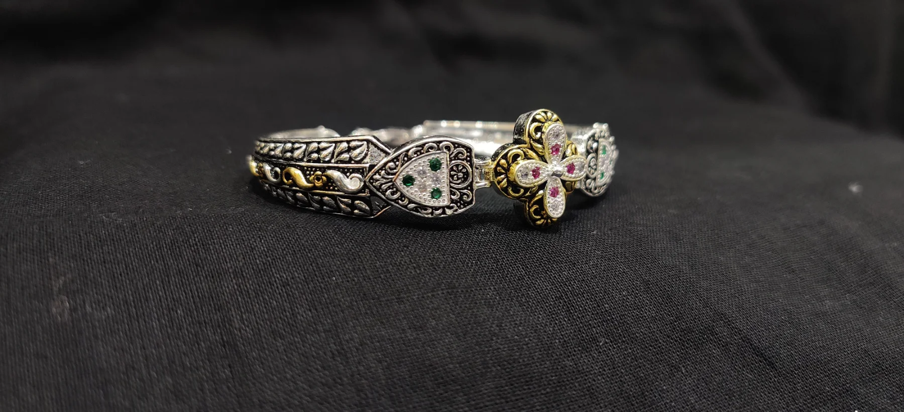 Oxidised silver bracelet featuring floral motifs, intricate patterns, and sparkling white, green, and red stones with gold polish touches.