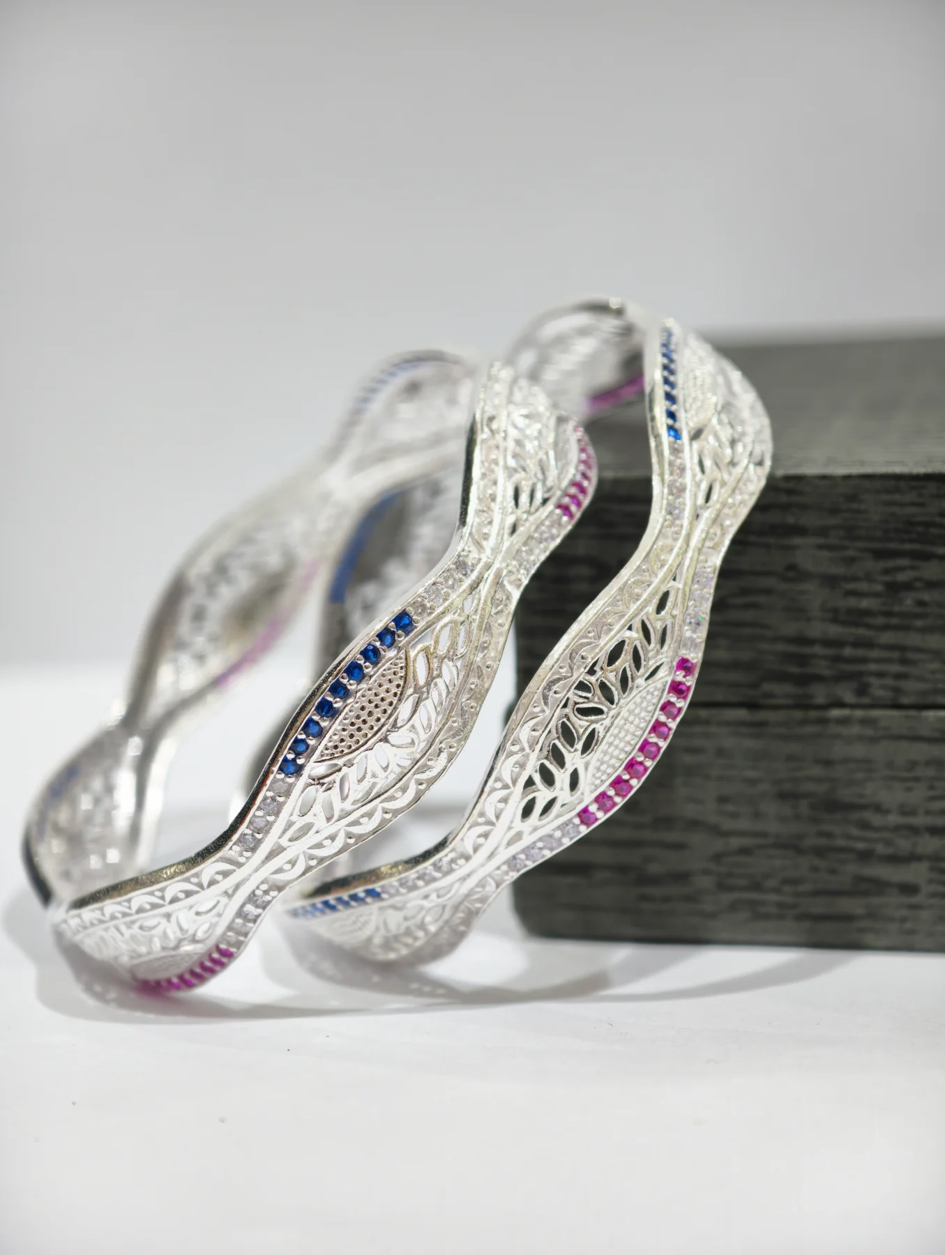 These wavy silver bangles feature intricate cutwork patterns accented with sparkling white, blue, and pink stones.