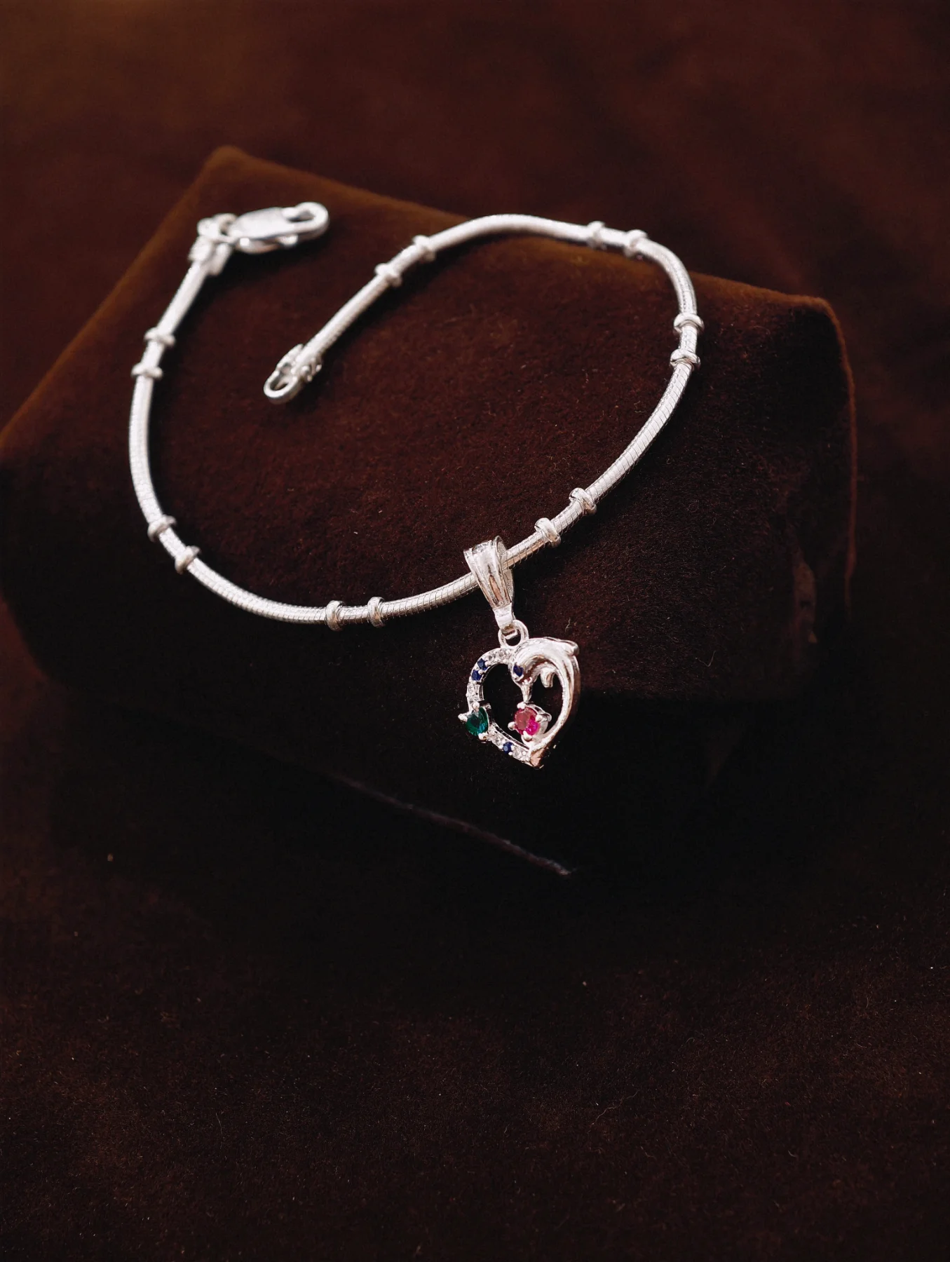 Silver chain bracelet with a heart shaped pendant featuring multicolored stones and a dolphin motif.