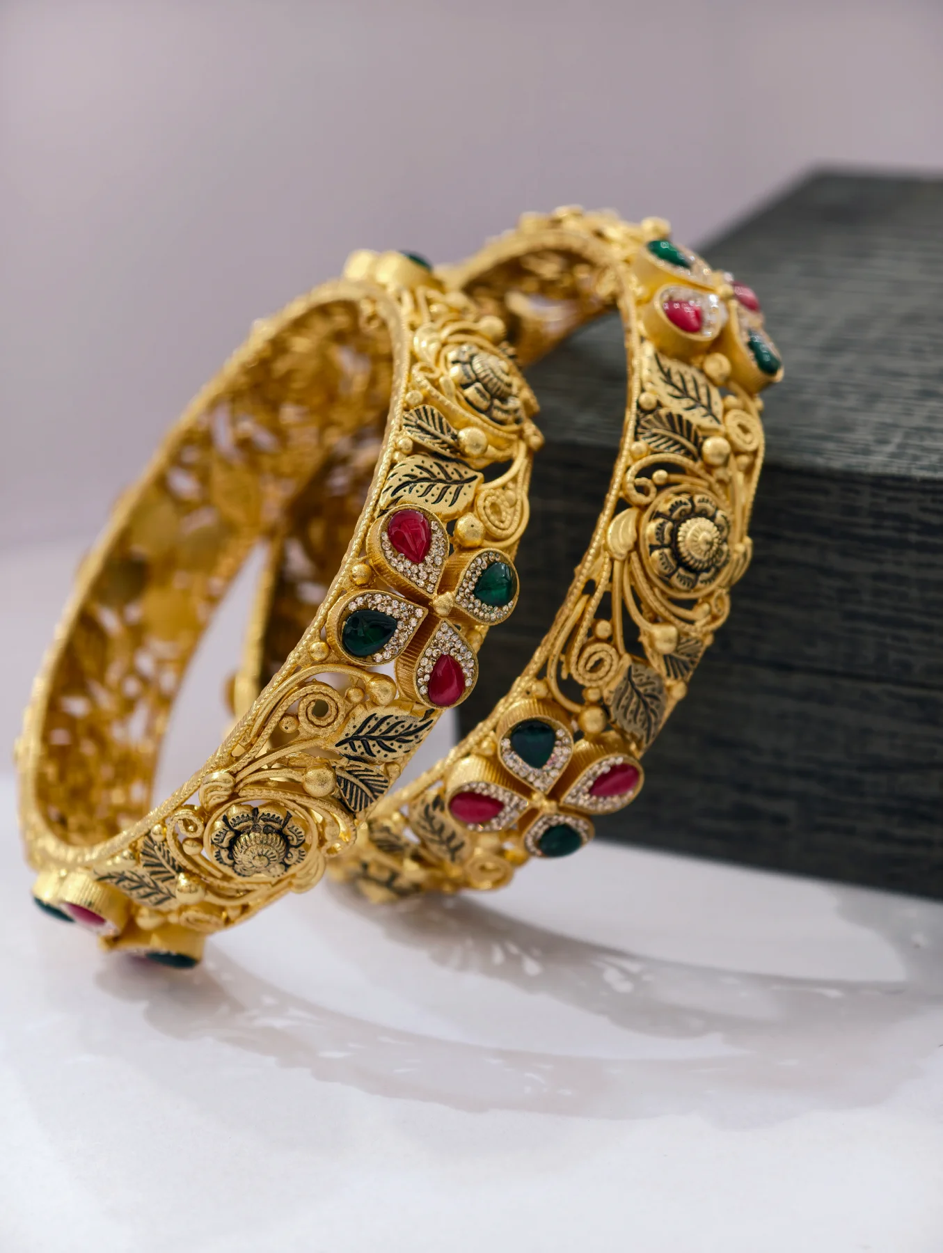Gold polished silver kadas featuring floral motifs, intricate filigree work, and embedded red and green stones.
