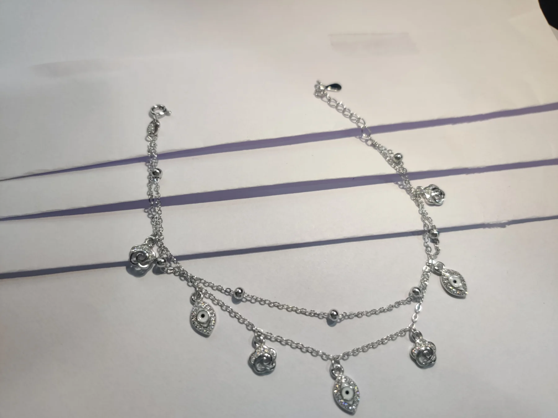 Delicate double chain silver bracelet featuring small dangling evil eye and flower charms with sparkling white stones.
