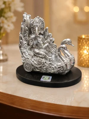 Glossy silver idol of Lord Ganesha sitting on an ornate swan chariot with intricate floral patterns and wheels.