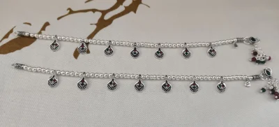 These elegant silver anklets feature a beaded chain with hanging pear shaped charms decorated with red and green stones.