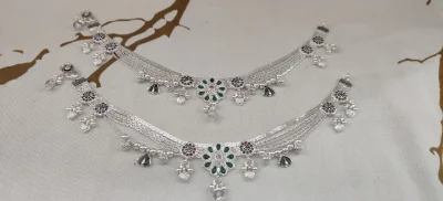 Fancy silver payal featuring multi strand chains, floral motifs with green and red stones, dangling beads, and ghungroos.