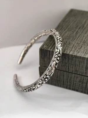 An oxidised silver kada with intricate floral and leaf carvings throughout the curved body.