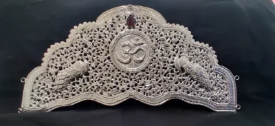 Silver crown featuring intricate filigree work, a central Om symbol, two peacocks, and a dark red stone.