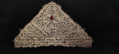 This triangular silver crown features intricate floral filigree work and a central red stone.