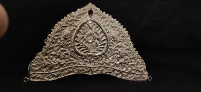 A triangular silver crown featuring detailed floral engraving, a central heart motif, and a small red stone at the top.