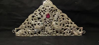 Silver crown featuring intricate jali work, peacock motifs, and a central red stone.