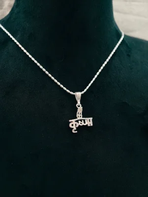 This silver chain features a textured pendant shaped like the Hindi word Krishna topped with a small leaf design.