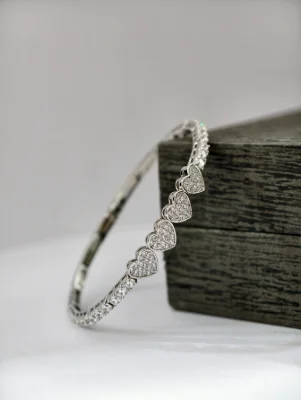 A silver kada bracelet featuring heart shaped motifs studded with small white stones along the band.