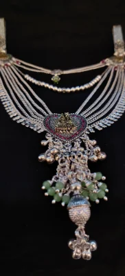 This multi layered silver waist chain features a central heart shaped motif with figures and dangling green beads and bells.