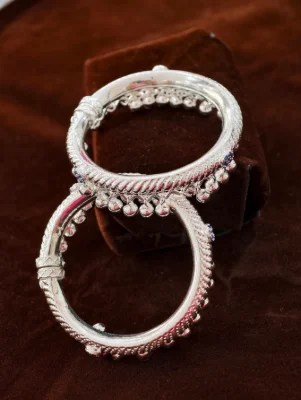 These circular children silver kada bangles feature a twisted rope pattern with hanging small silver beads and blue stone accents.