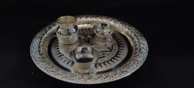 This shiny silver worship thali features intricate floral and sunray patterns, complete with a small matching pot and two bowls.