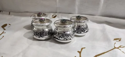 Polished silver dishes featuring intricate floral engravings, rounded bodies, and lids adorned with central coloured stones.