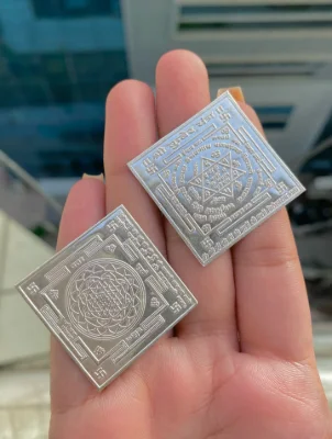Two square silver religious yantras featuring intricate engraved geometric patterns and auspicious symbols.