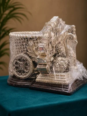 An ornate silver decorative plaque shaped like a chariot with intricate peacock and wheel motifs.