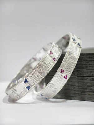 A pair of silver bangles with a textured surface and decorative floral motifs inlaid with red, blue and white stones.